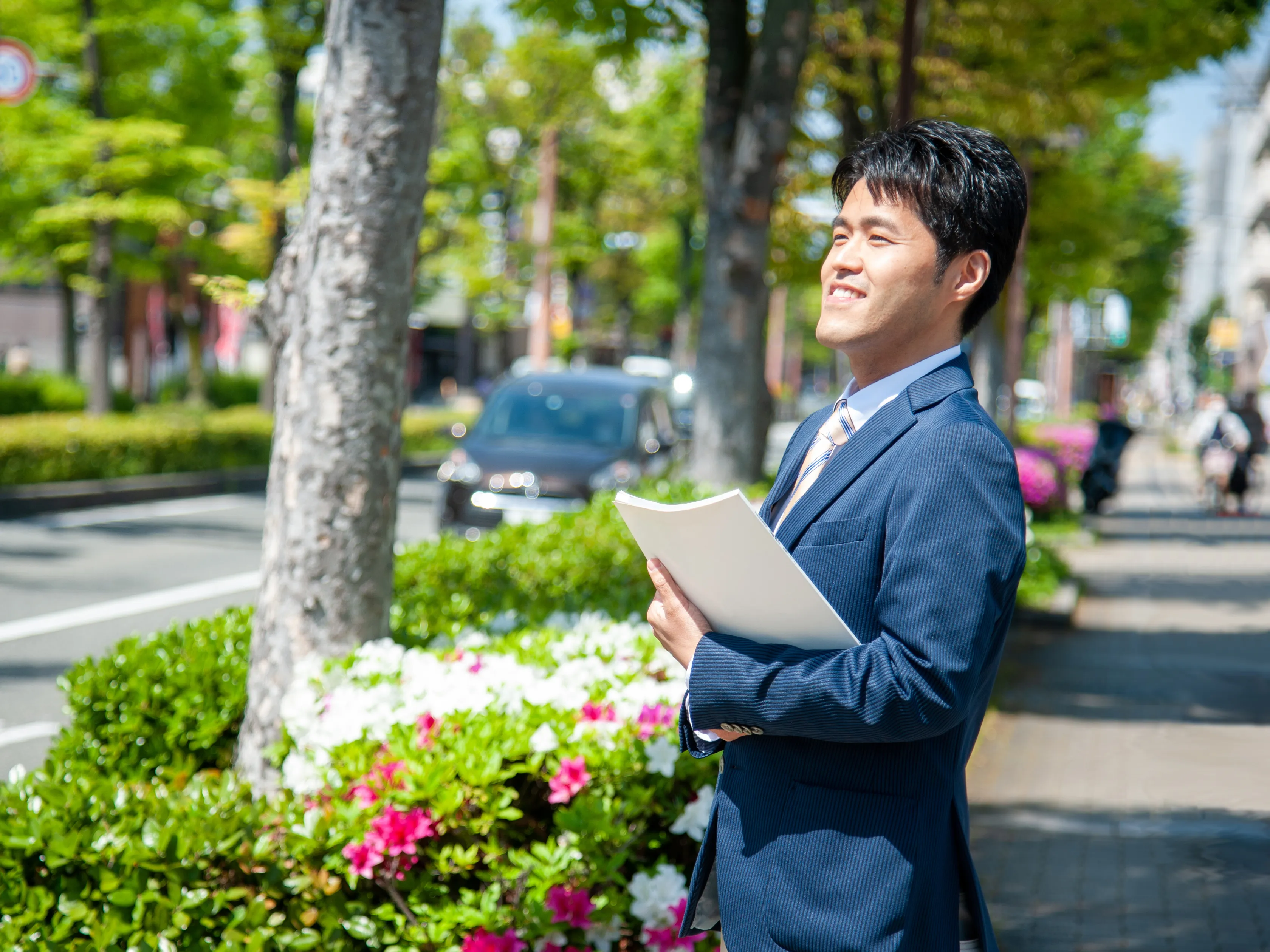 写真:つつじの咲く街路樹のある歩道を背景に、紺色のスーツを着た黒髪の男性が手に書類を持って微笑んでいる